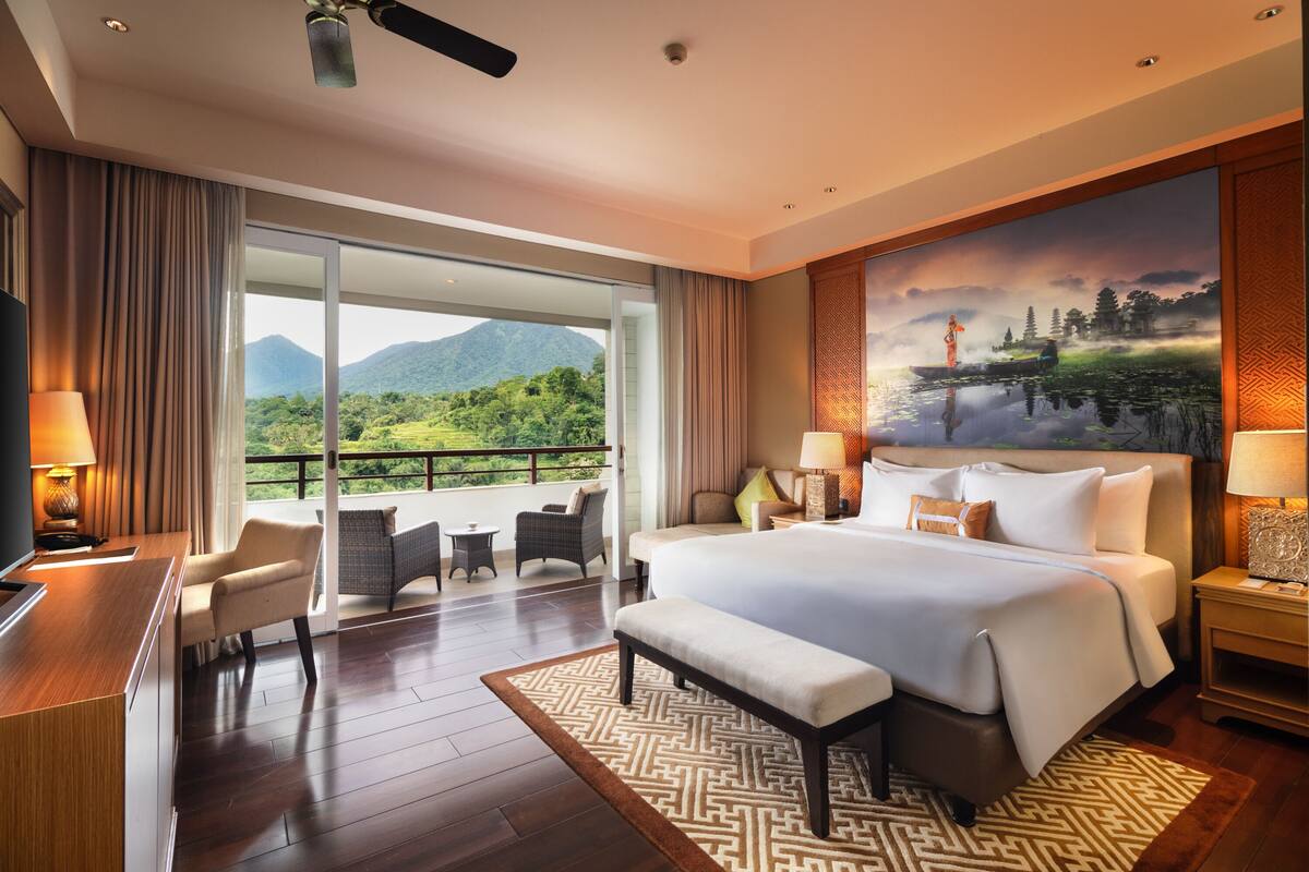 A hotel room with a large bed, a desk, a chair, a bench, and a view of a mountainous landscape.