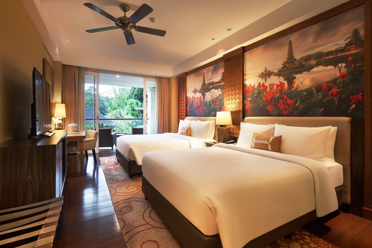A hotel room with two beds, a desk, a chair, a ceiling fan, and a large window with a view of greenery.