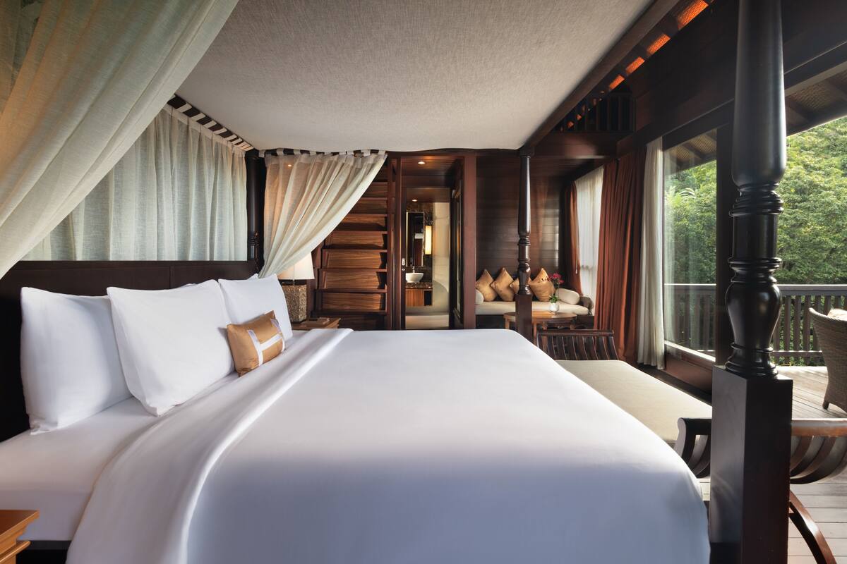 A large bed with white linens, a wooden headboard, and a view of a balcony with wicker furniture.