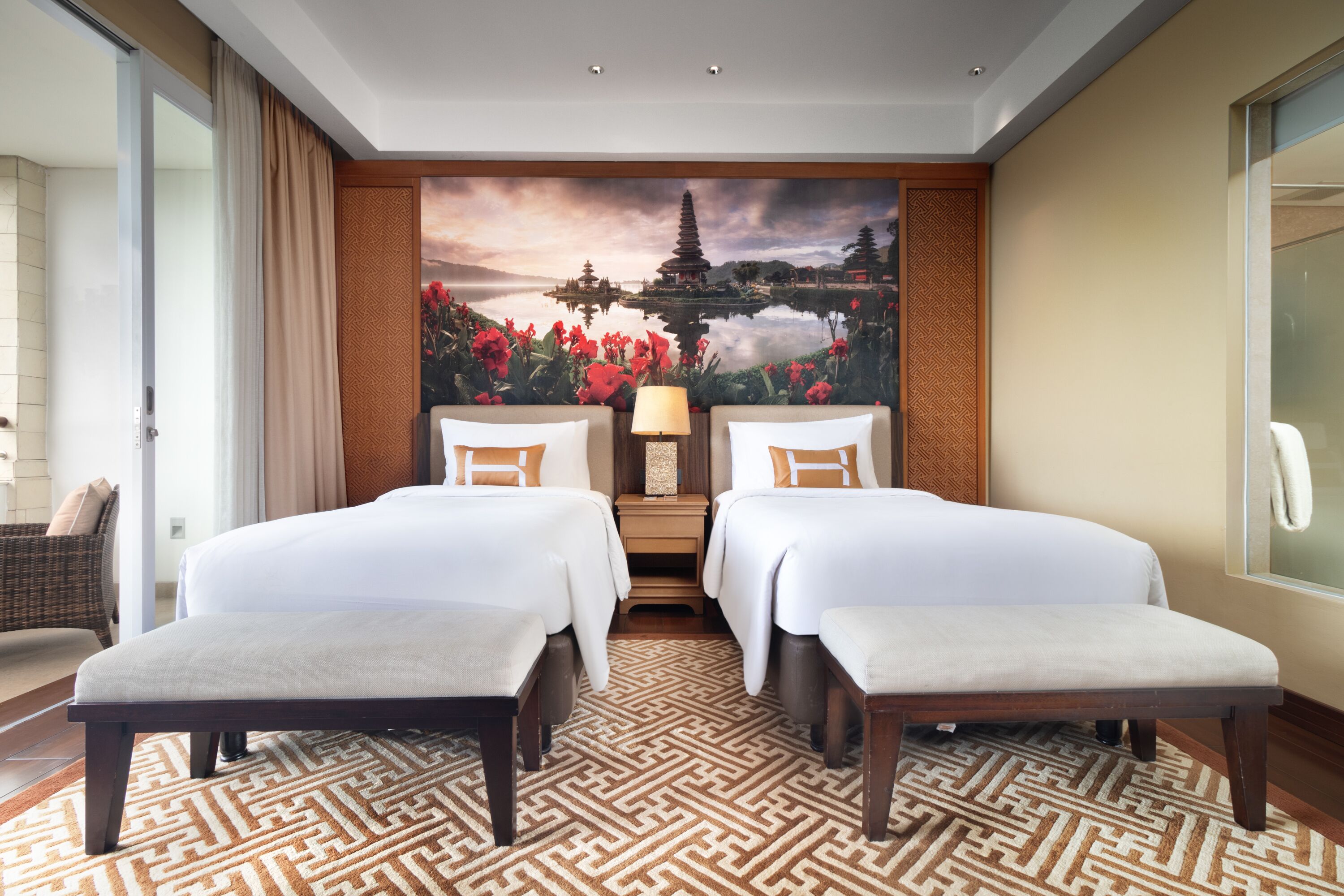 A hotel room with two beds, a large painting of a temple and water, a wooden nightstand, and a patterned rug.