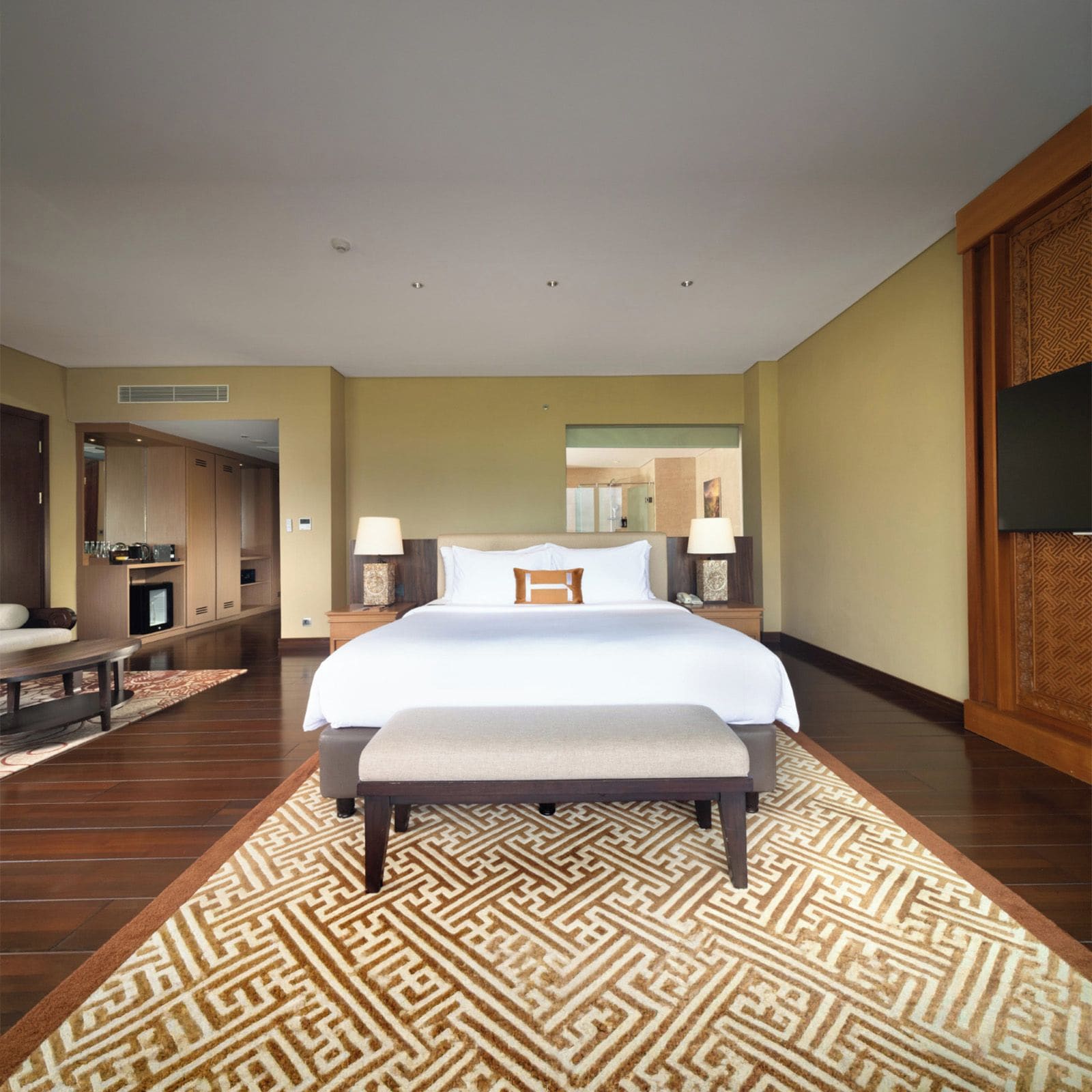 Quarto de hotel com cama grande, luminárias de cabeceira, um banco, televisão e tapete estampado.