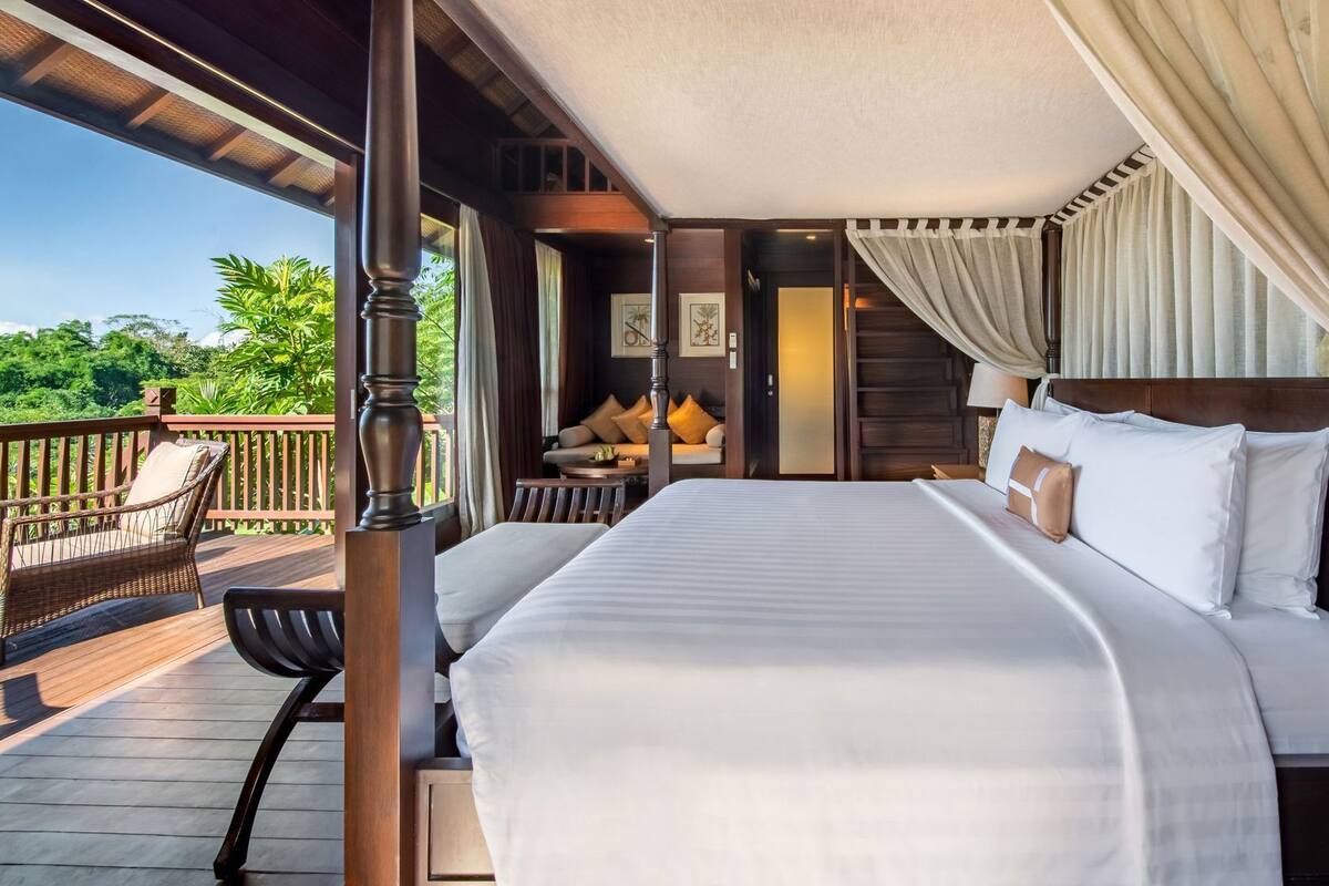 A hotel room with a large bed, a wooden headboard, a balcony with a chair, and a view of greenery.