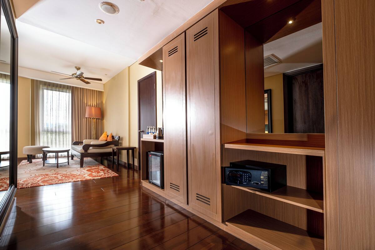 A hotel room with wooden flooring, a ceiling fan, a seating area with a table and chairs, a minibar, and a large wardrobe with built-in shelves.