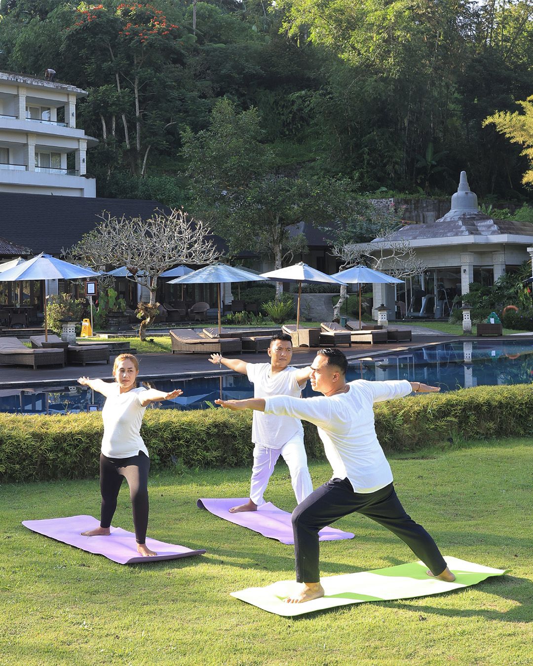 A group of people practicing yoga outdoors with a pool and umbrellas in the background.
