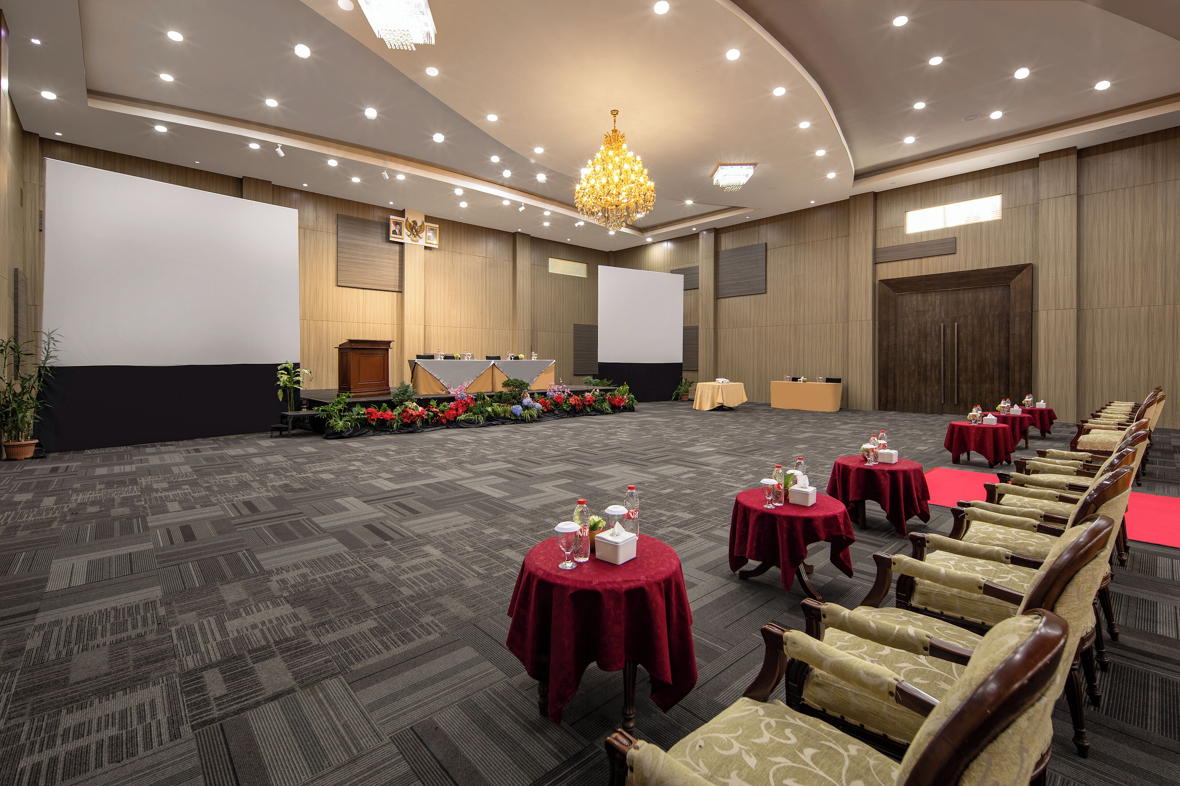 A conference room with a stage, rows of chairs, tables with red tablecloths, and a chandelier.