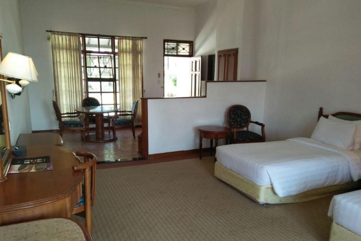 A hotel room with a bed, a desk with a lamp, a chair, a small table, and a window with curtains.