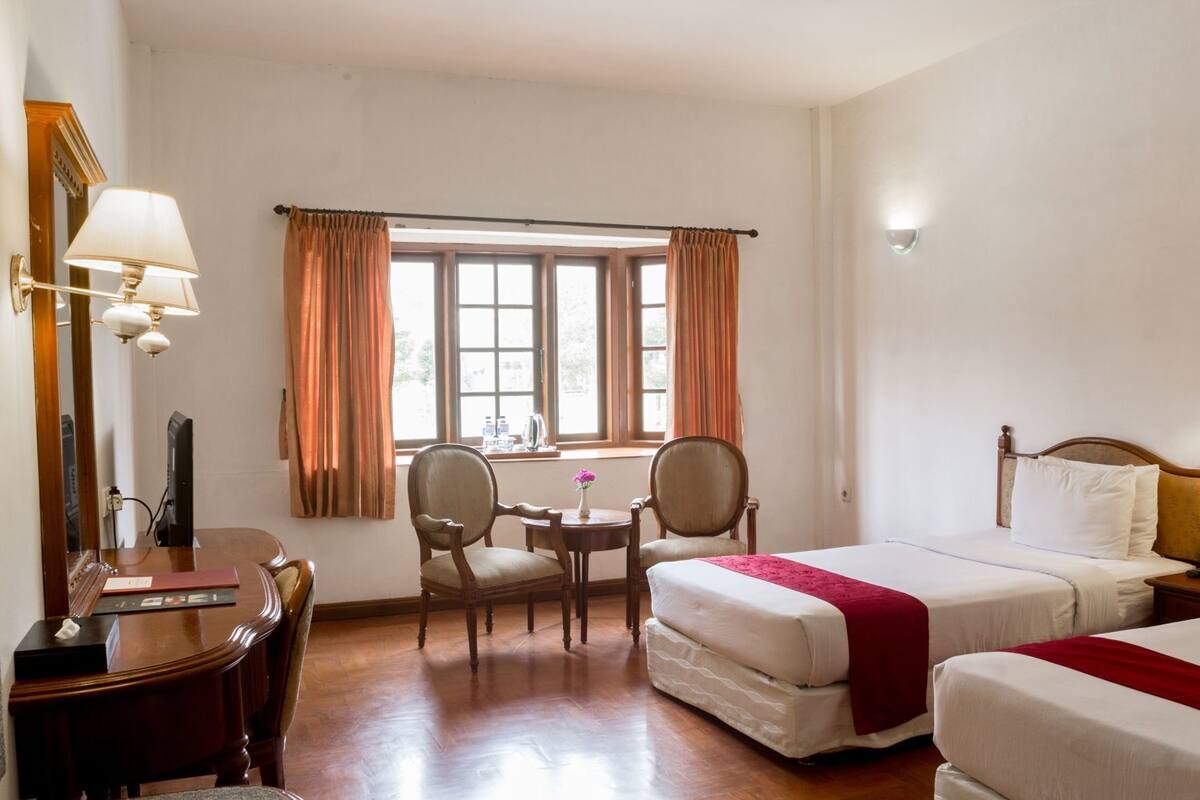 Una habitación de hotel con una cama, dos sillas, un escritorio, una lámpara y un ventanal con cortinas.