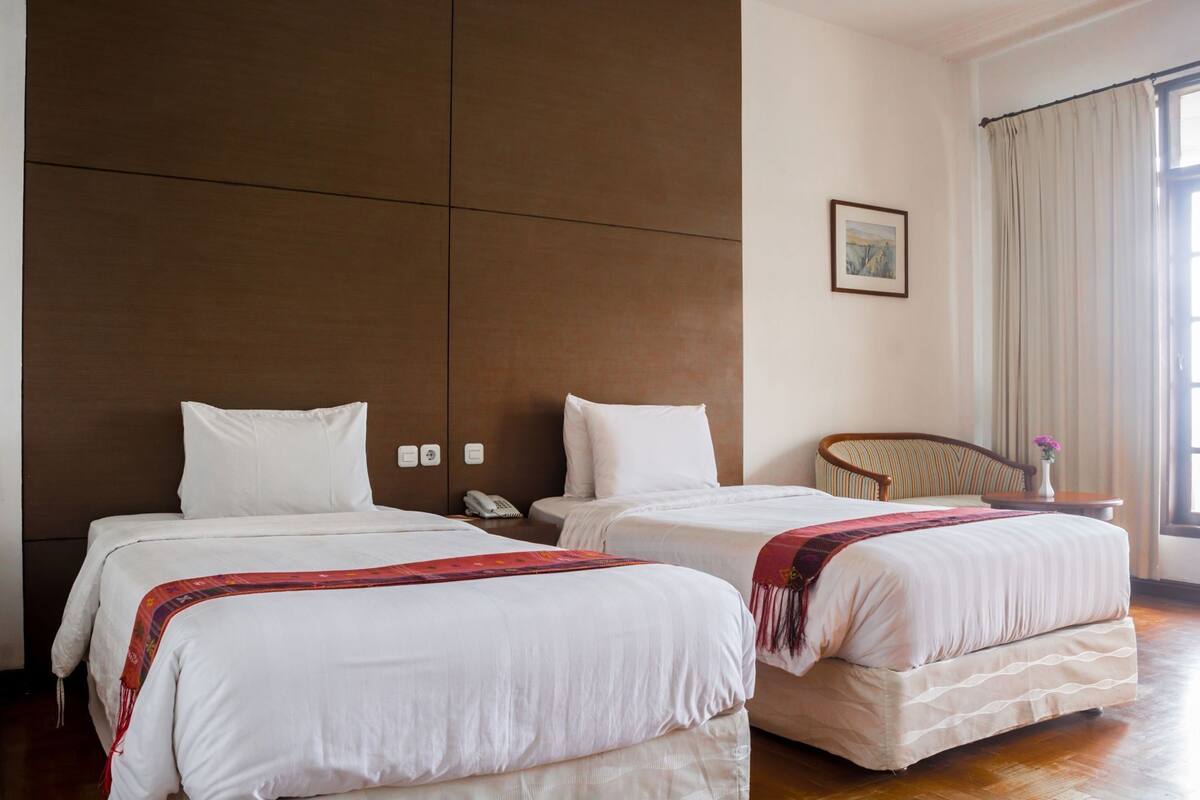 Habitación de hotel con dos camas, cabecero de madera, teléfono, una silla y ventana con cortinas.