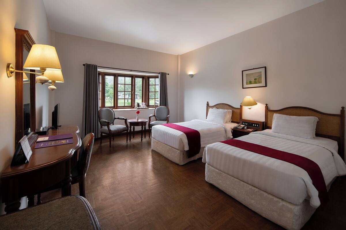 Habitación de hotel con dos camas, un escritorio, una silla, una mesa y un ventanal con cortinas.