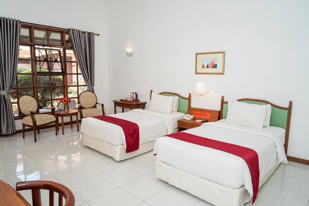 Habitación de hotel con dos camas, un rincón de descanso con sillas, una mesita y una ventana con cortinas.