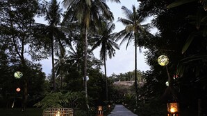 A pathway lined with palm trees and lanterns, leading to a structure with a thatched roof. - Hotel Tugu Lombok (Tanjung)
