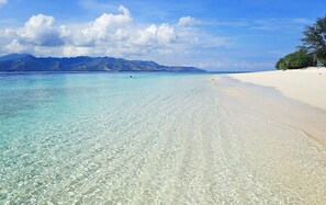 Una playa con aguas turquesas cristalinas, arena blanca y montañas al fondo.