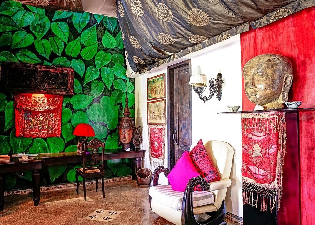A room with a green leafy wall, a red curtain, a Buddha statue, and a traditional chair.
