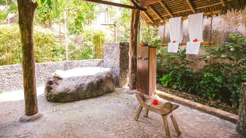A stone bathtub under a wooden pergola with a thatched roof, surrounded by lush greenery.