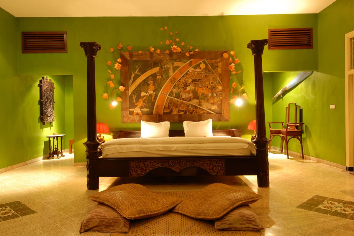 Een hemelbed met een gedetailleerd hoofdeinde, een tafeltje met een lamp en een stoel in een kamer met groene accenten.