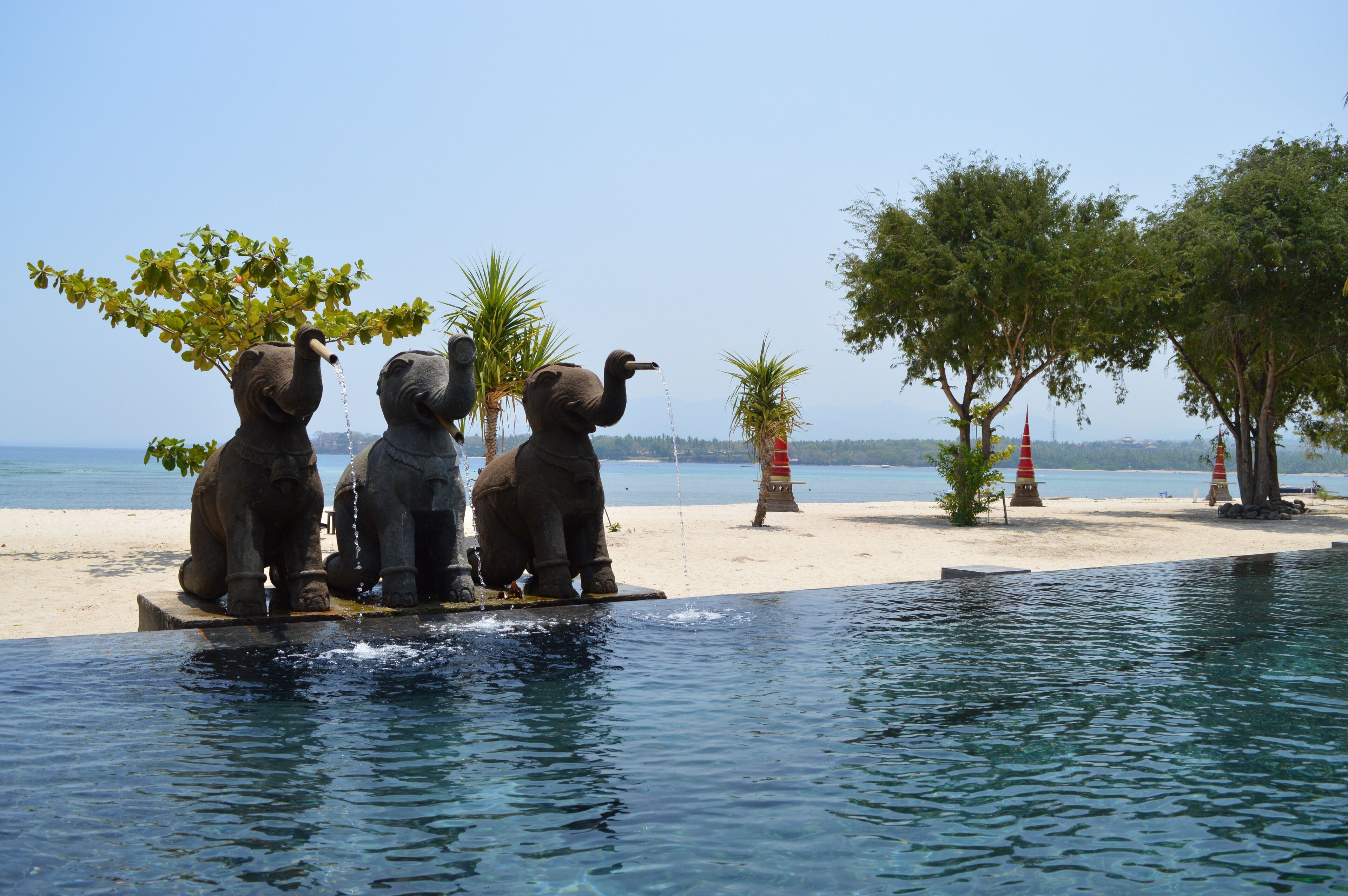 Un área de piscina con tres estatuas de elefante, una playa arenosa y árboles al fondo.