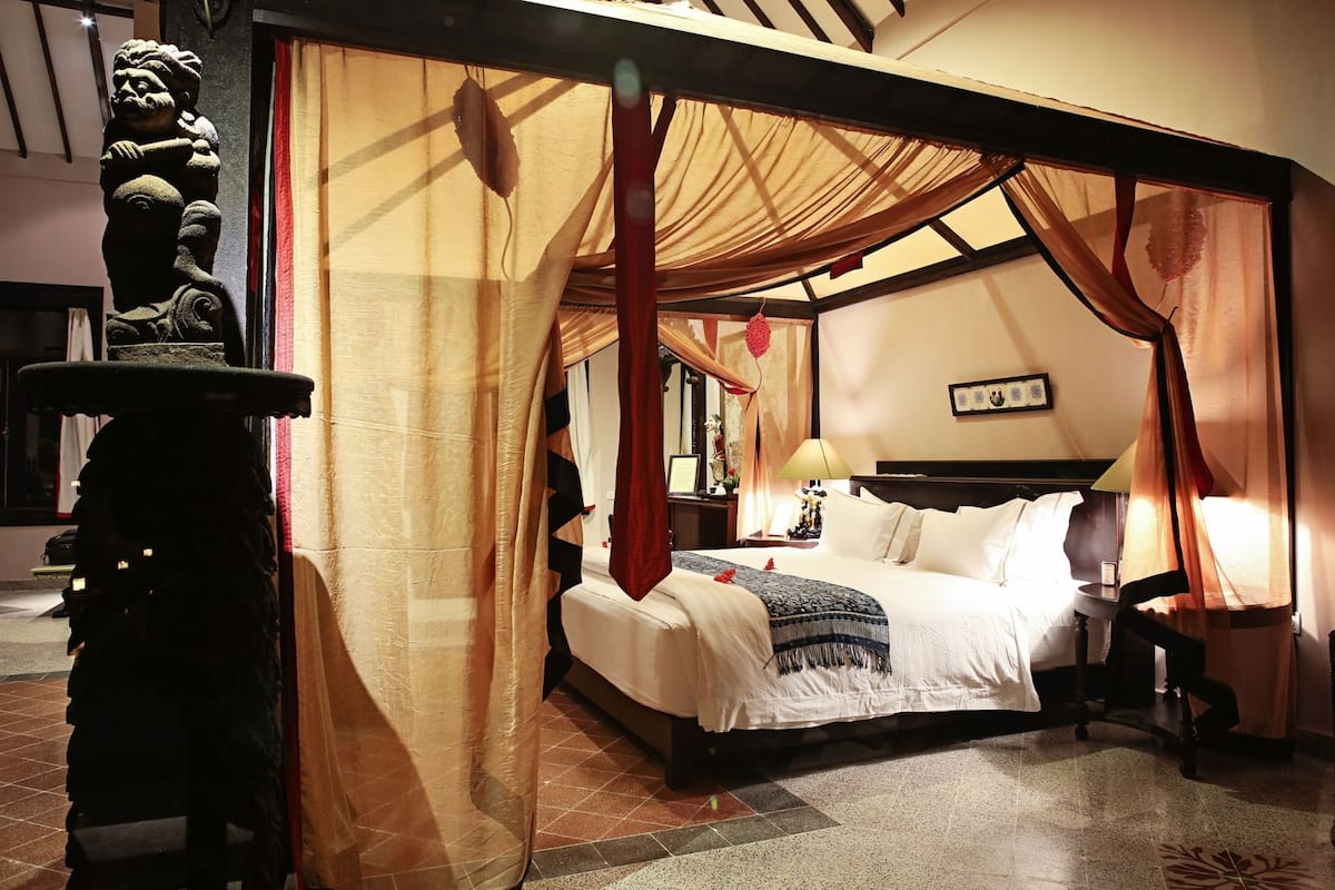 Een hemelbed met baldakijn, een nachtkastje met lamp en een beeld in de hoek.