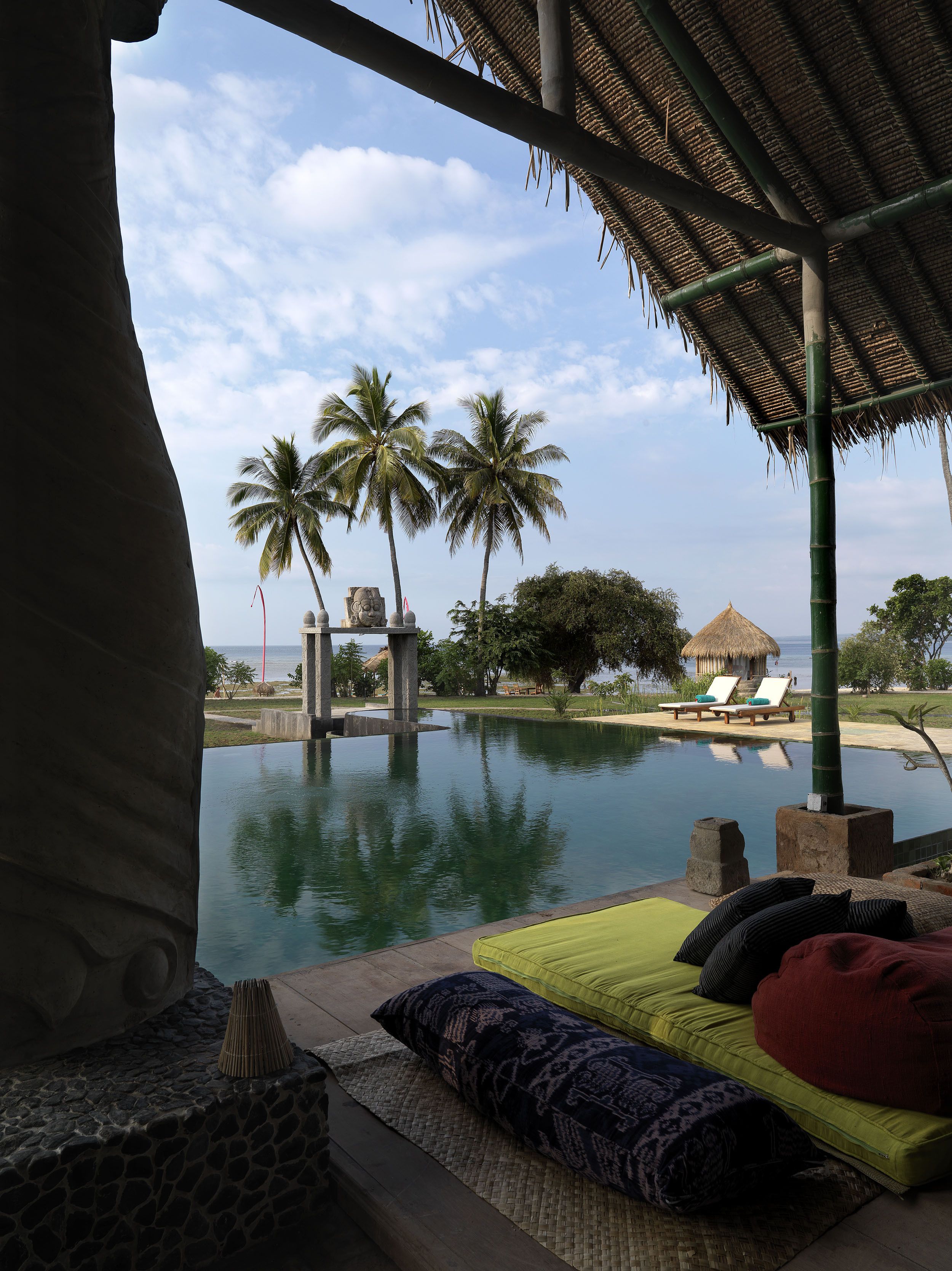 Un espace aménagé au bord de la piscine, avec des chaises longues, une structure dotée d’un toit de chaume, des palmiers et une vue sur la mer.