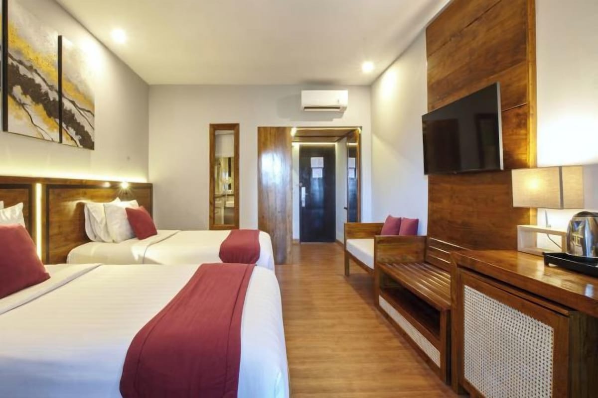 A hotel room with two beds, a wooden floor, a TV, and a desk with a lamp.