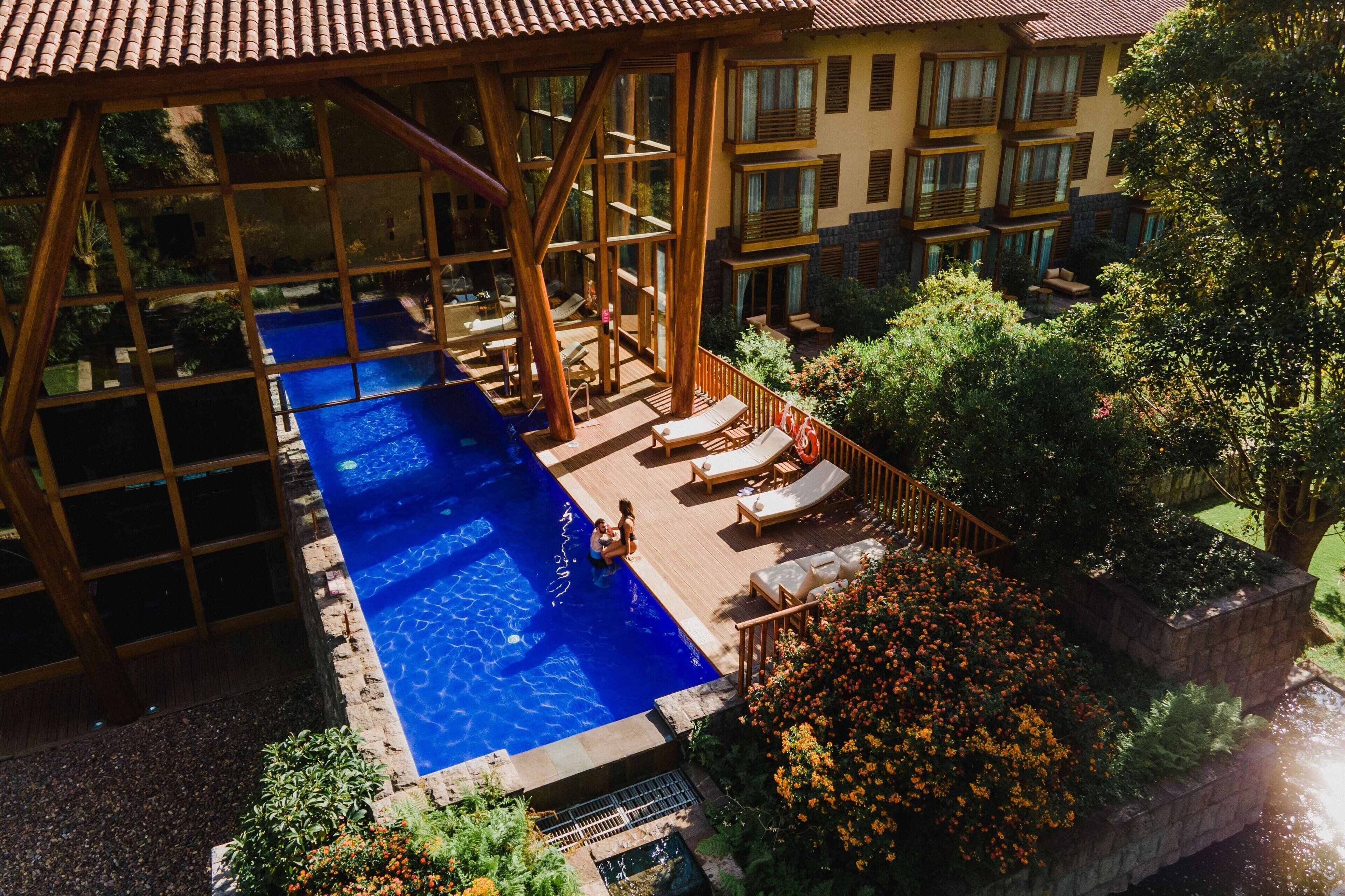 An outdoor swimming pool with a wooden deck and lounge chairs, surrounded by greenery and a building with multiple balconies.