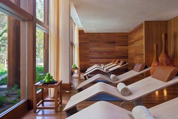 Un cuarto de spa con paredes de madera y ventanas amplias, varias camillas de masaje y una pequeña mesa con manzanas.