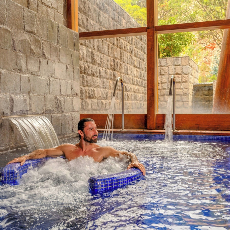 A man in a hot tub with a waterfall feature, stone walls, and large windows.