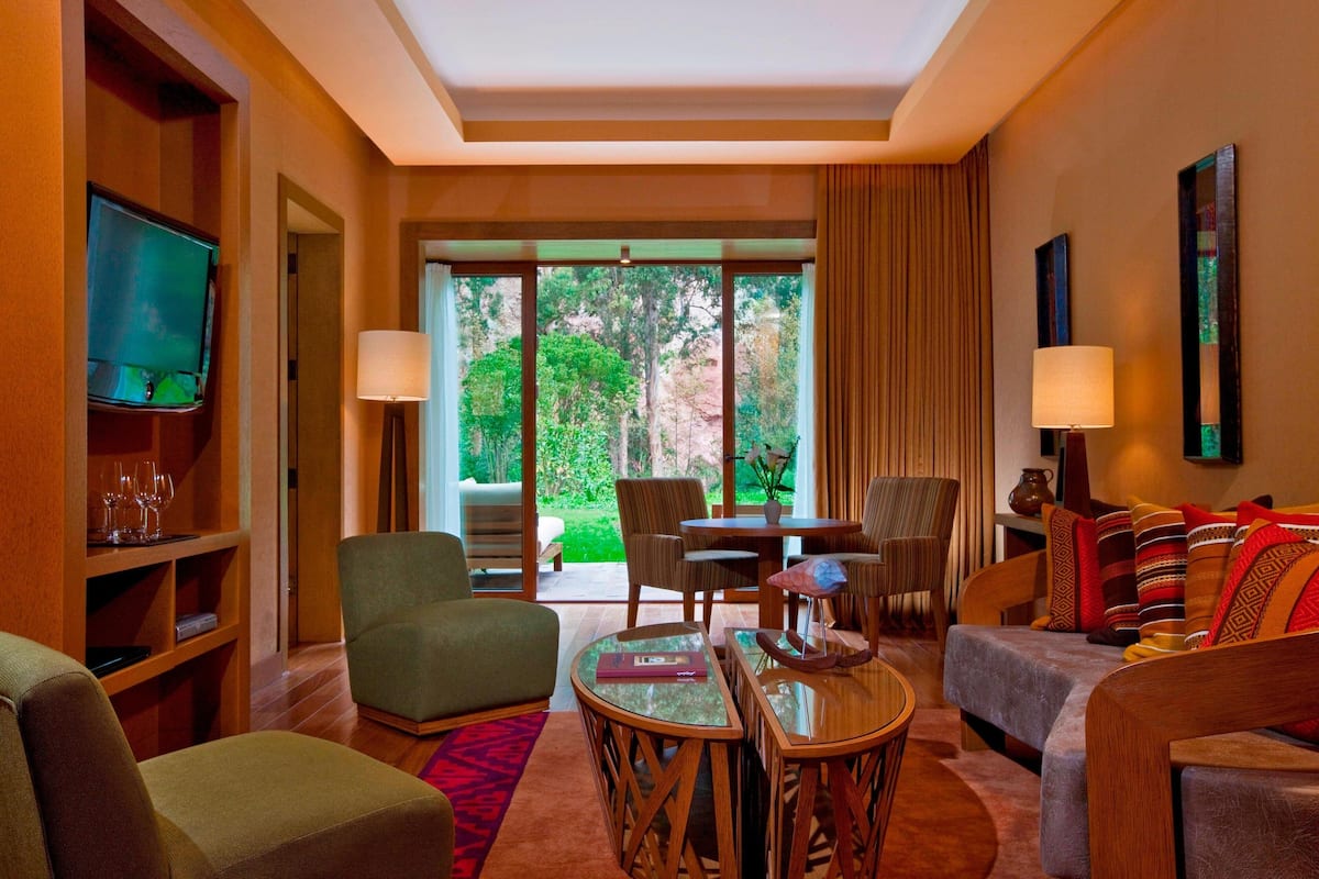 A hotel room with a sofa, armchair, dining table, and chairs. There is a television on the left and a window with curtains.