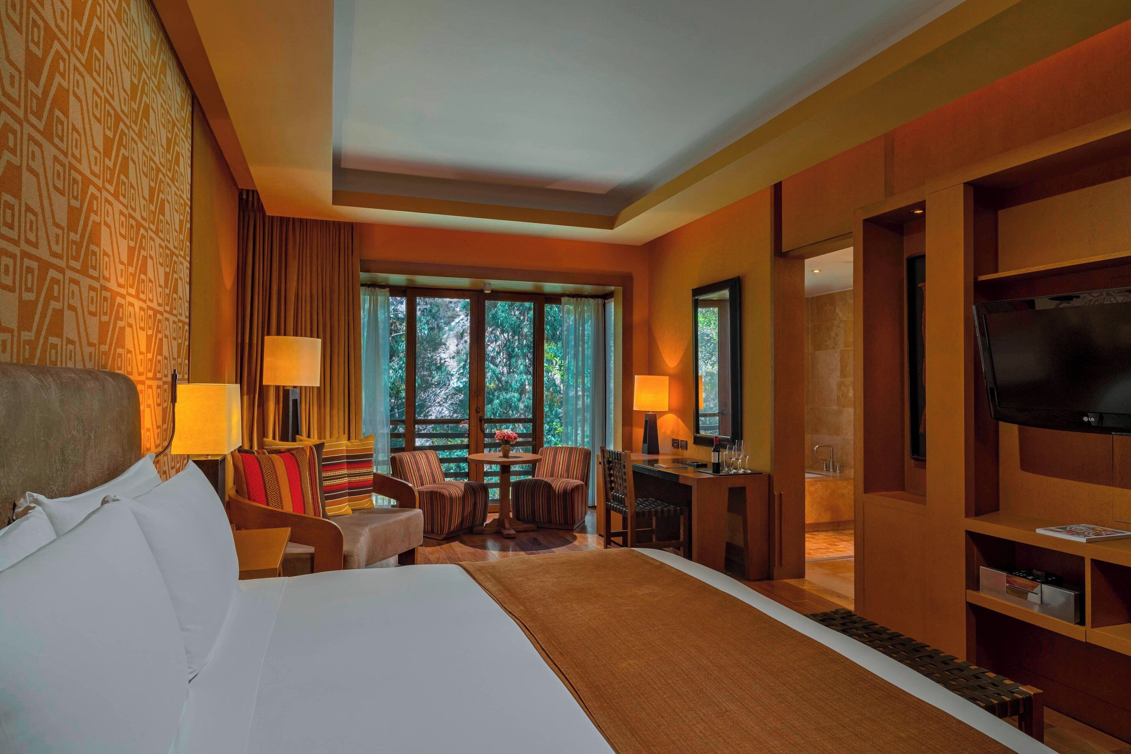 A hotel room with a large bed, a desk, a TV, and a view of trees through the window.