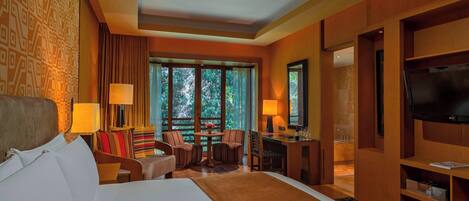 A hotel room with a large bed, a desk, a TV, and a view of trees through the window.