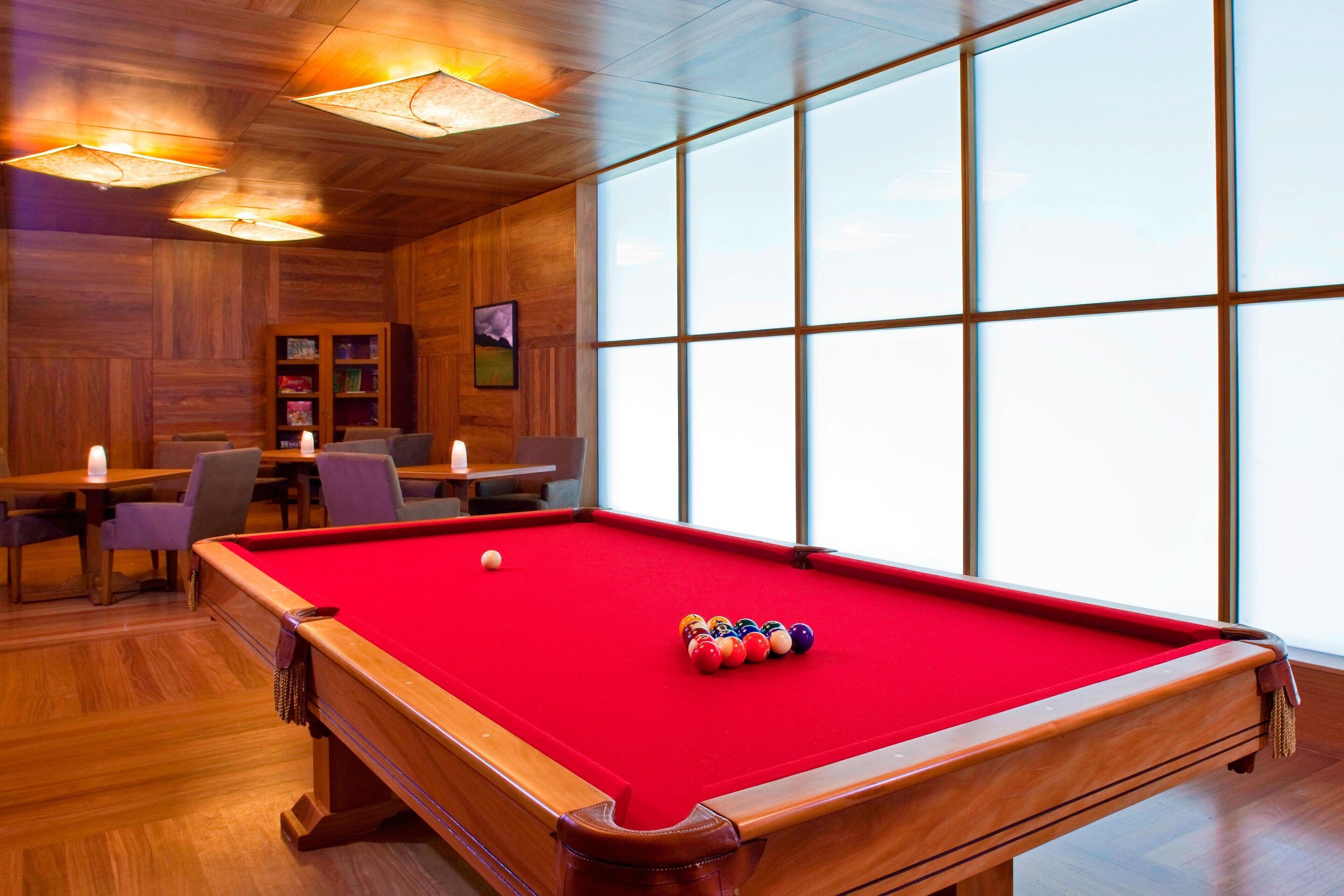A pool table with a red felt surface, wooden walls, and large windows.