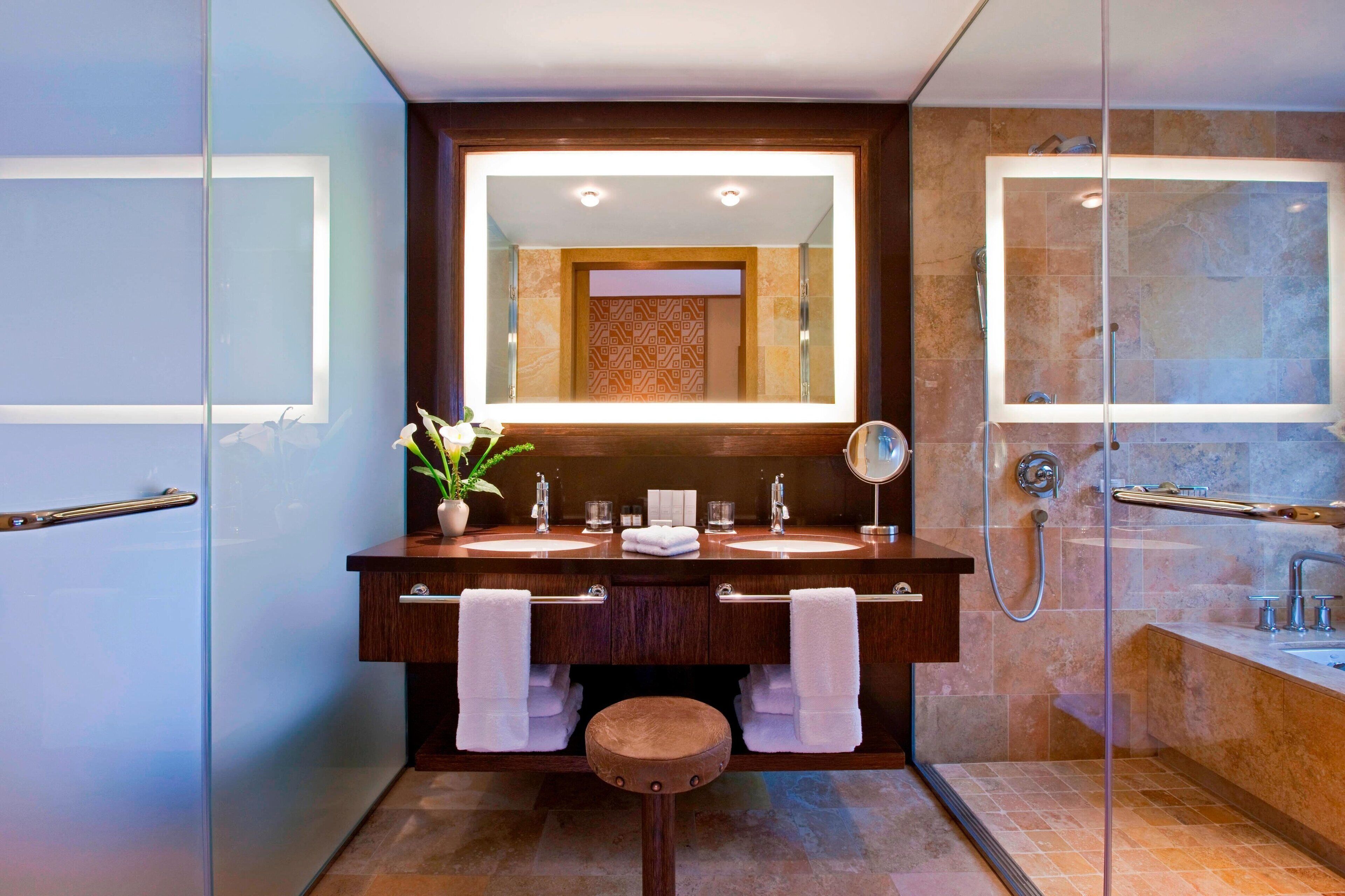 Un baño moderno con dos lavamanos, un espejo grande, zona de ducha y un taburete de madera.