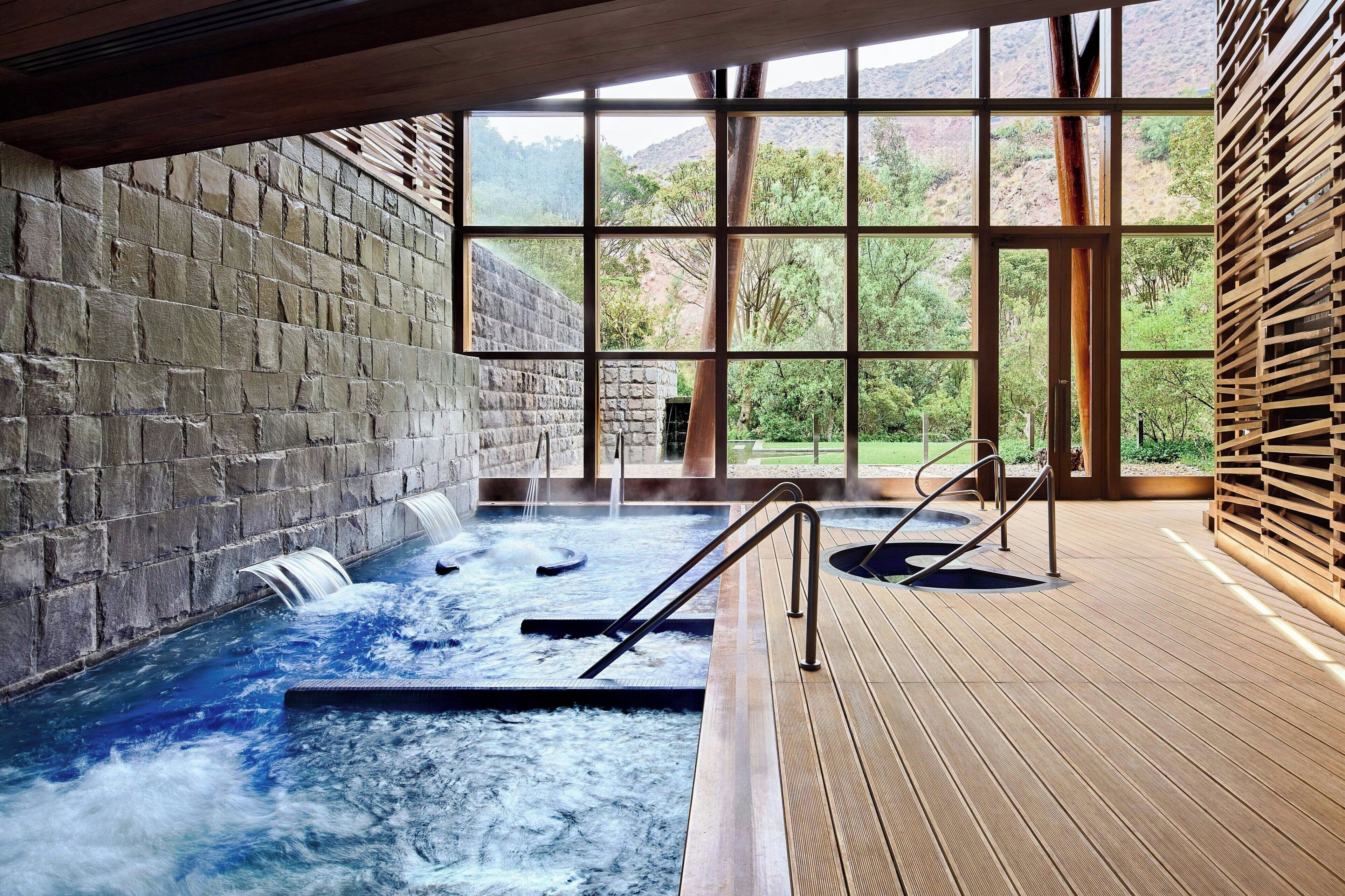 Un área de spa con jacuzzi, terraza de madera, paredes de piedra y ventanas amplias con vistas a la vegetación.