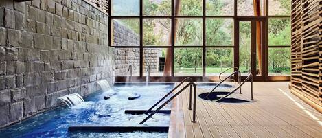 Un área de spa con jacuzzi, terraza de madera, paredes de piedra y ventanas amplias con vistas a la vegetación.