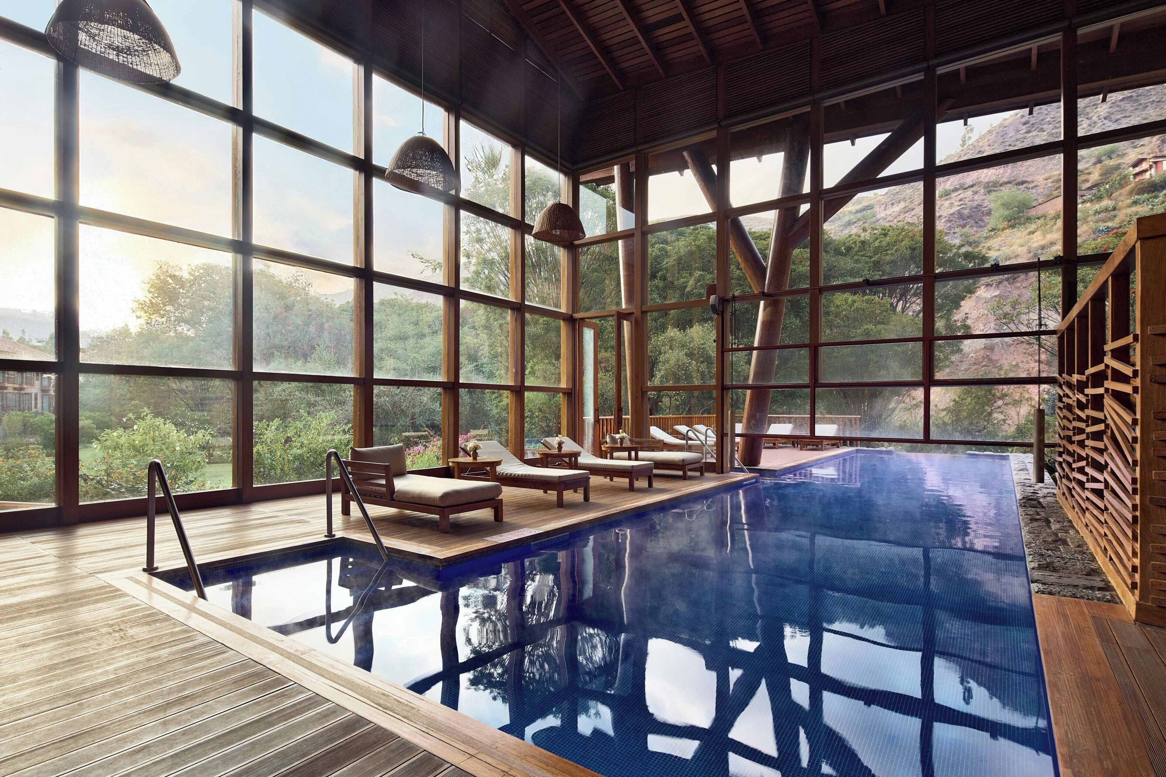 A large indoor swimming pool with wooden deck, lounge chairs, and large windows offering a view of a mountainous landscape.
