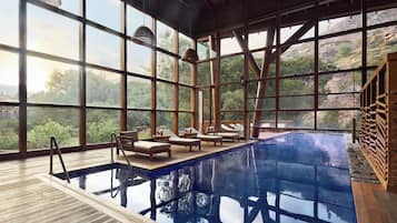 A large indoor swimming pool with wooden deck, lounge chairs, and large windows offering a view of a mountainous landscape.