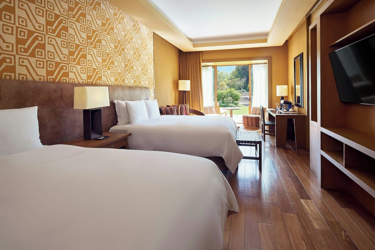 A hotel room with two beds, a desk, a TV, and a view of the outdoors.