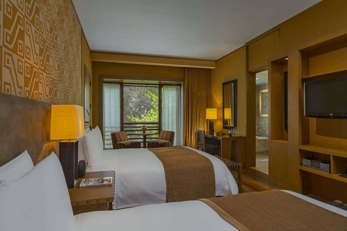 A hotel room with two beds, a desk, a TV, and a window with a view of greenery.