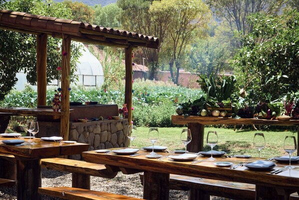 An outdoor dining area with wooden tables and benches, stone walls, and a greenhouse in the background.