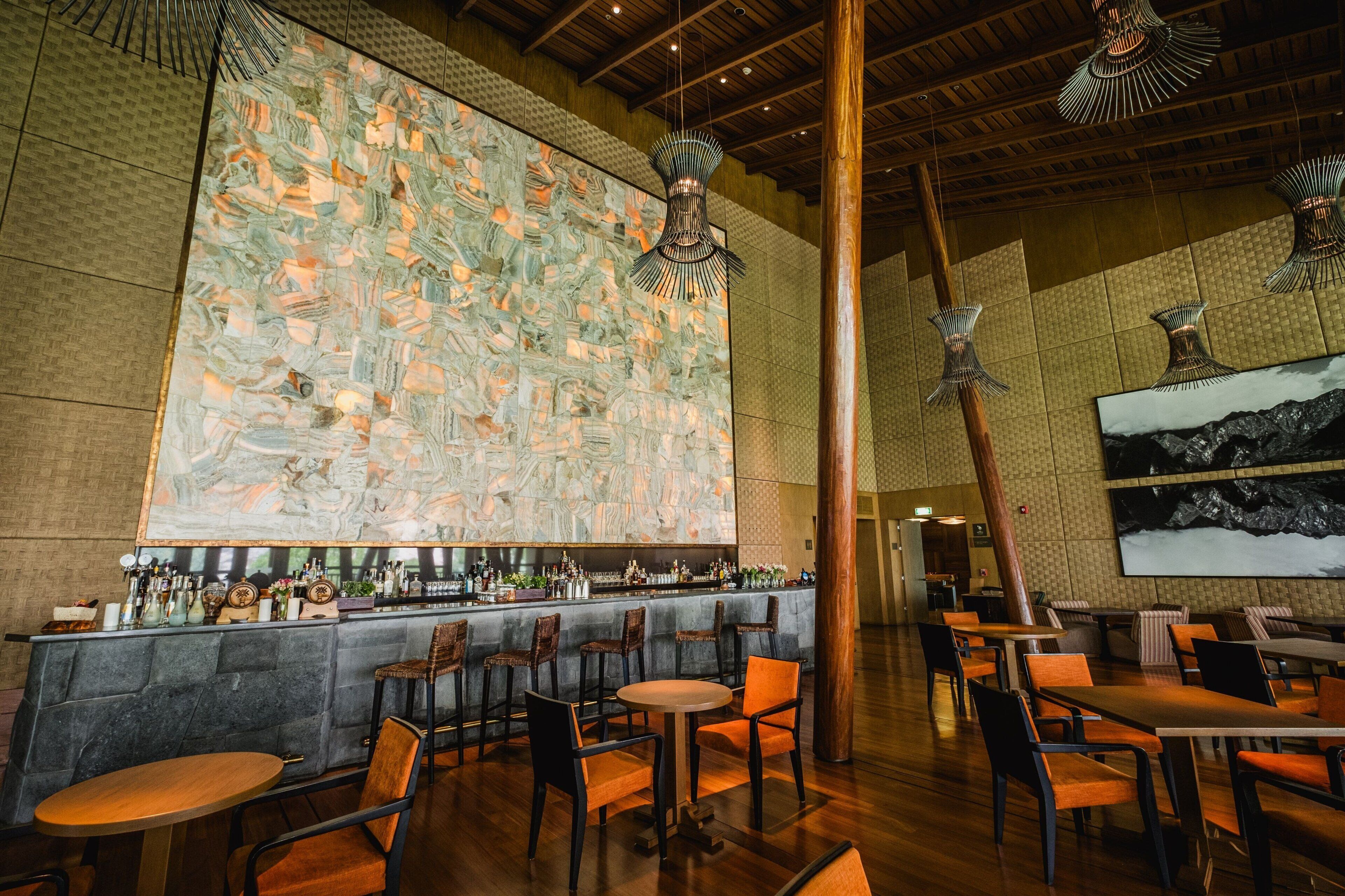 A bar area with a high ceiling, wooden beams, and hanging light fixtures. There are tables and chairs arranged for seating.
