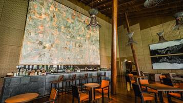 A bar area with a high ceiling, wooden beams, and hanging light fixtures. There are tables and chairs arranged for seating.