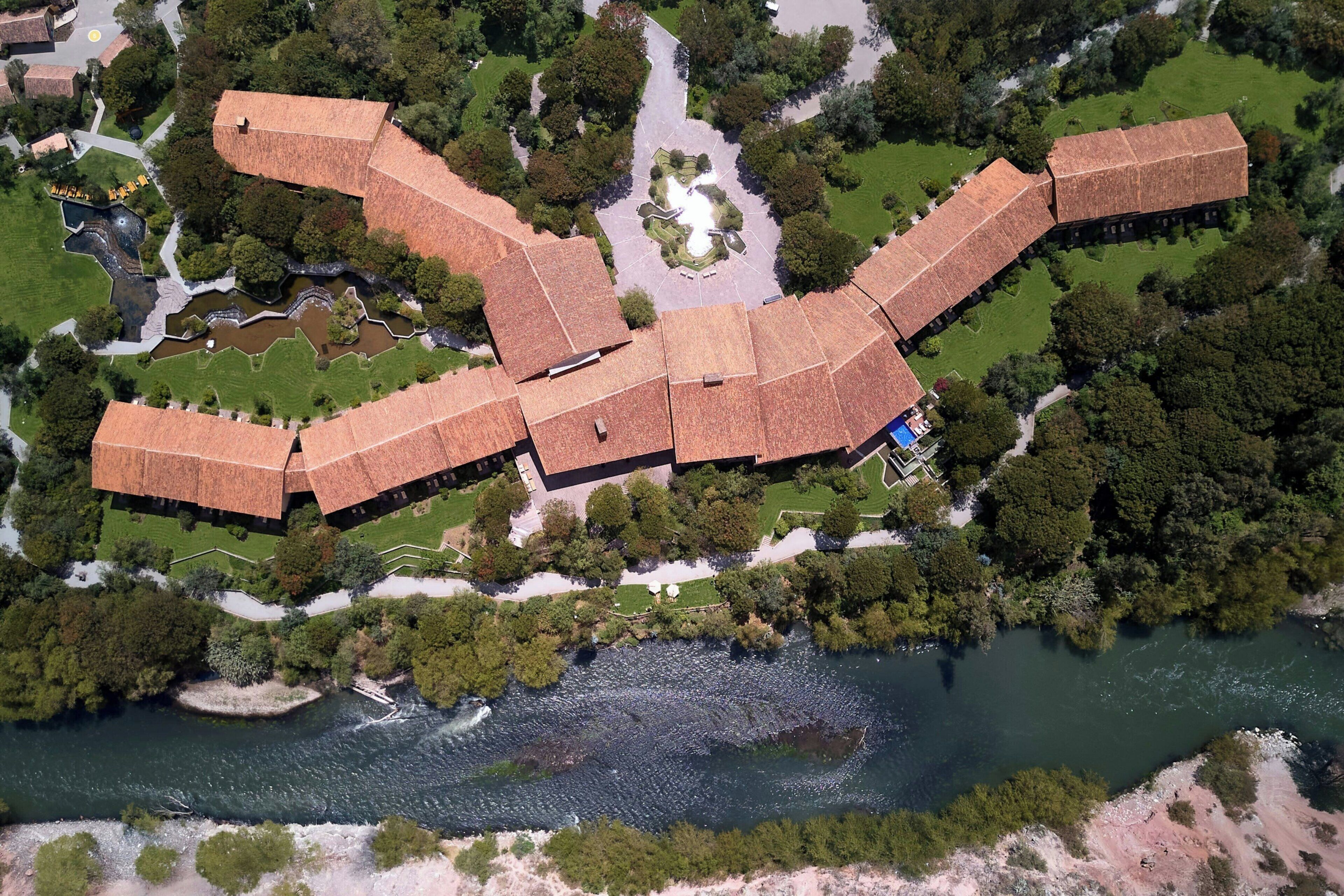 An aerial view of a hotel with a distinctive roof structure, surrounded by greenery and a river.
