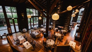 Un restaurant spacieux avec de hauts plafonds, des poutres en bois et de grandes fenêtres offrant une vue sur l’extérieur. Les tables sont disposées avec soin et recouvertes de nappes blanches, accompagnées de chaises.