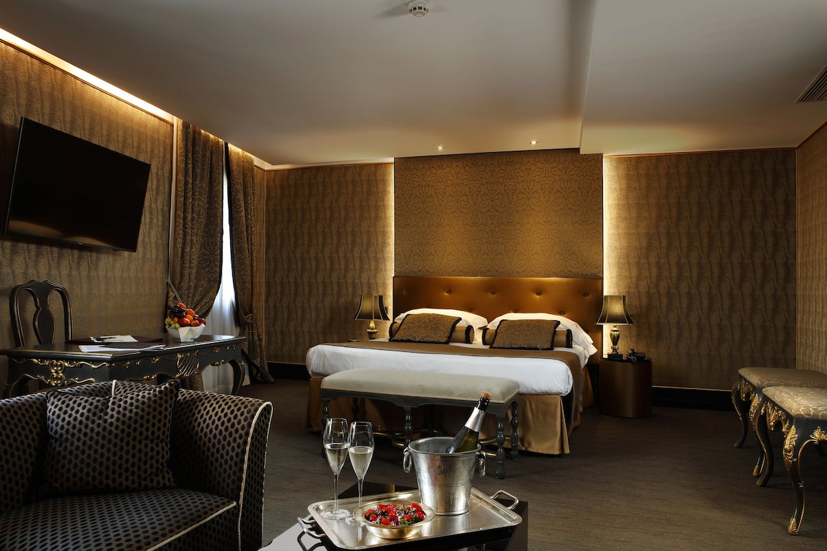 A hotel room with a bed, bedside tables, a TV, a dining area with a table and chairs, and a minibar.