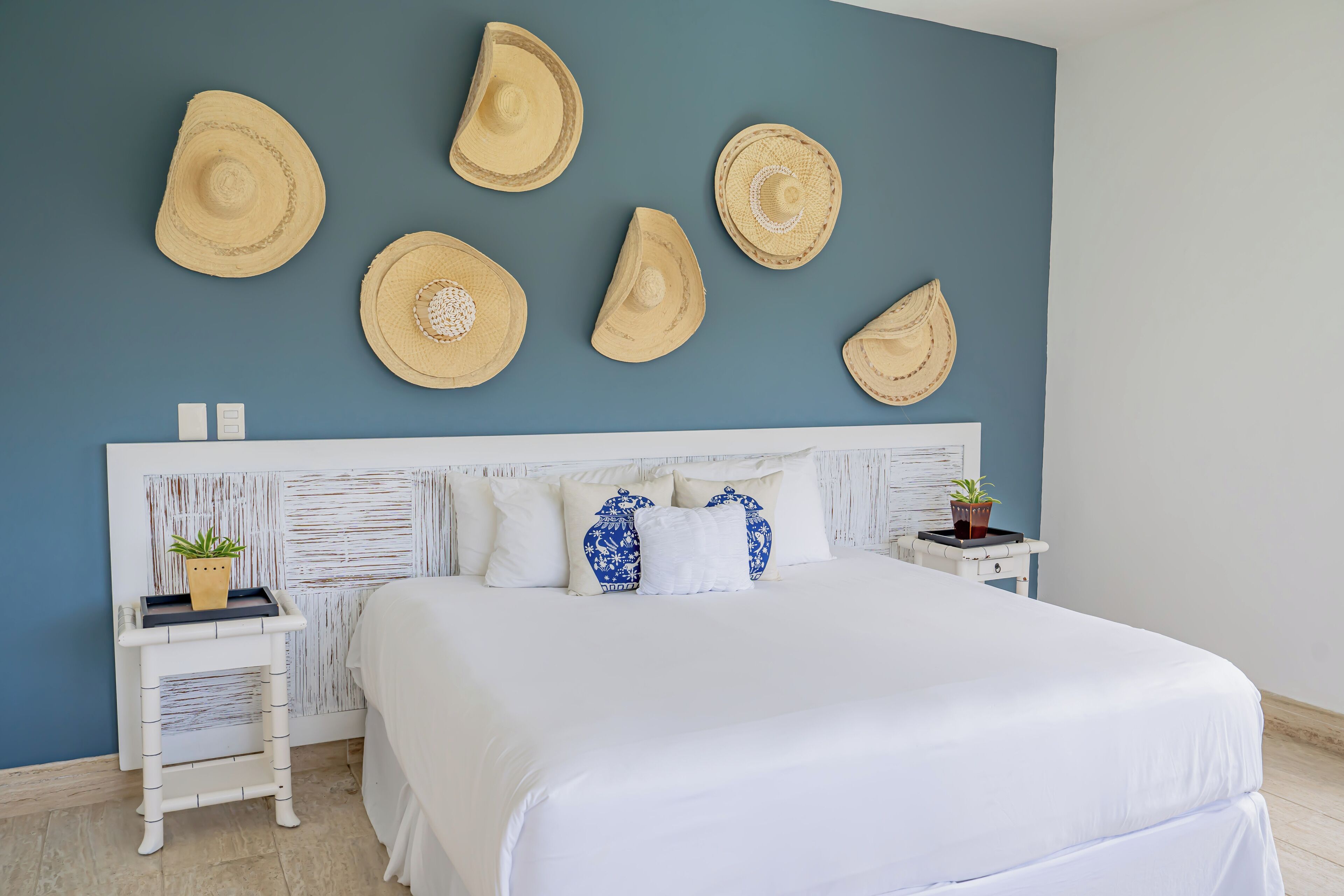 Een keurig opgemaakt bed met wit beddengoed, een hoofdbord met een reliëfpatroon, en een muur met verschillende hoeden. Er zijn twee nachtkastjes, waarvan één met een plantje en de andere met een klein potplantje.