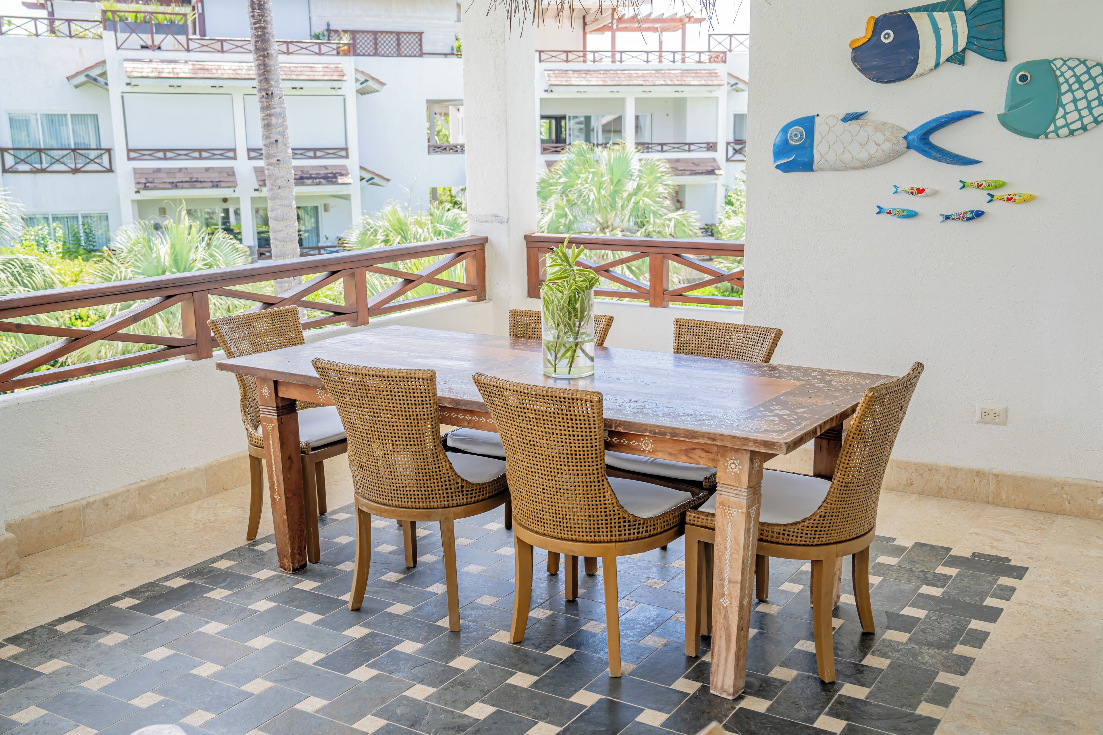 Un coin repas avec une table en bois et des chaises en osier, une plante en pot et une vue sur un immeuble doté de balcons.