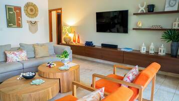 Een moderne woonkamer met een grijze bank, oranje fauteuils, houten salontafels en een aan de muur bevestigde televisie.