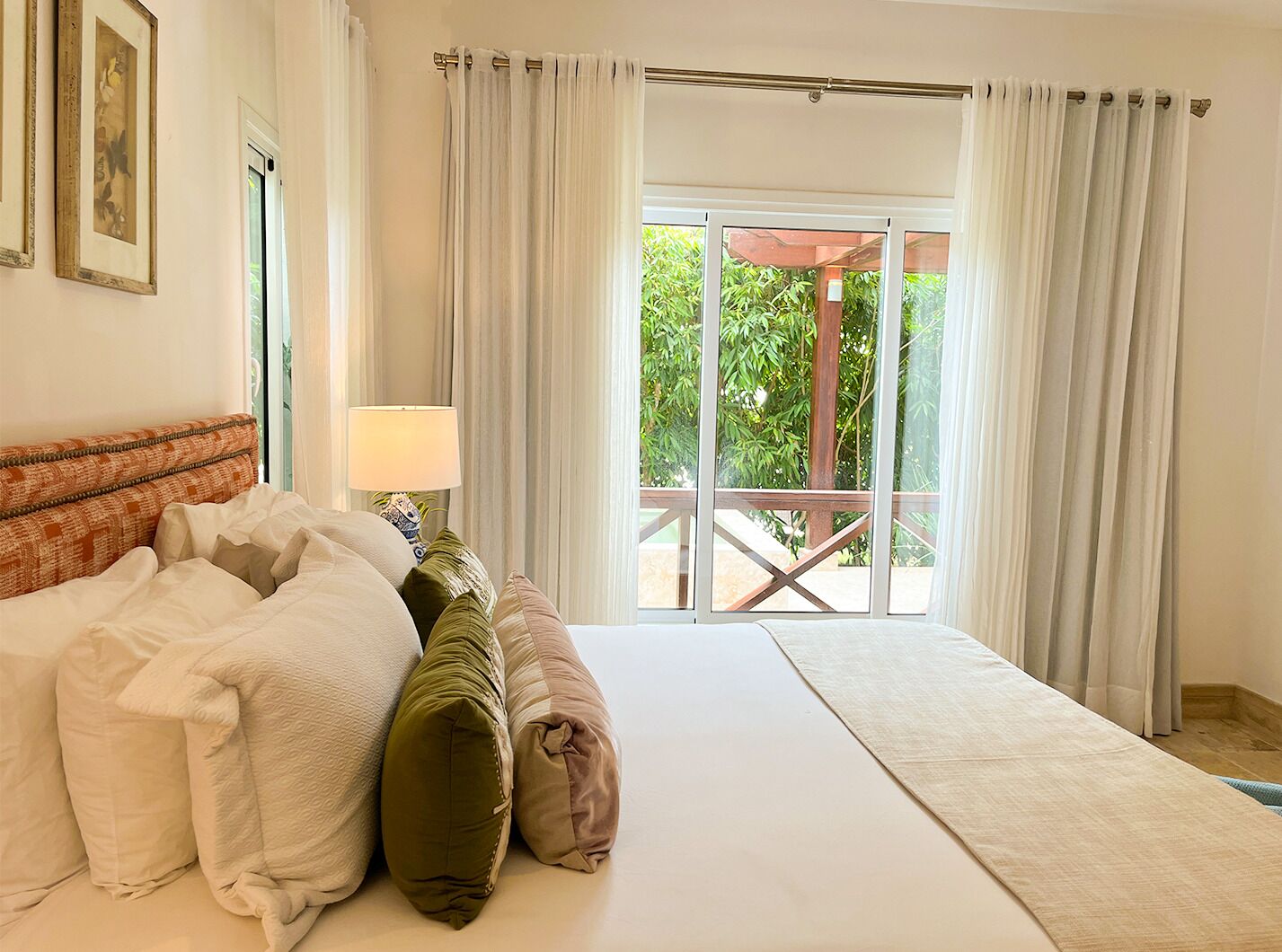 Een slaapkamer met een bed, kussens, een nachtkastje, een lamp en uitzicht op groen.