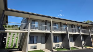 Un immeuble moderne à plusieurs étages, doté de balcons, d’un toit plat et d’un petit espace jardin aménagé avec des chaises et des tables.