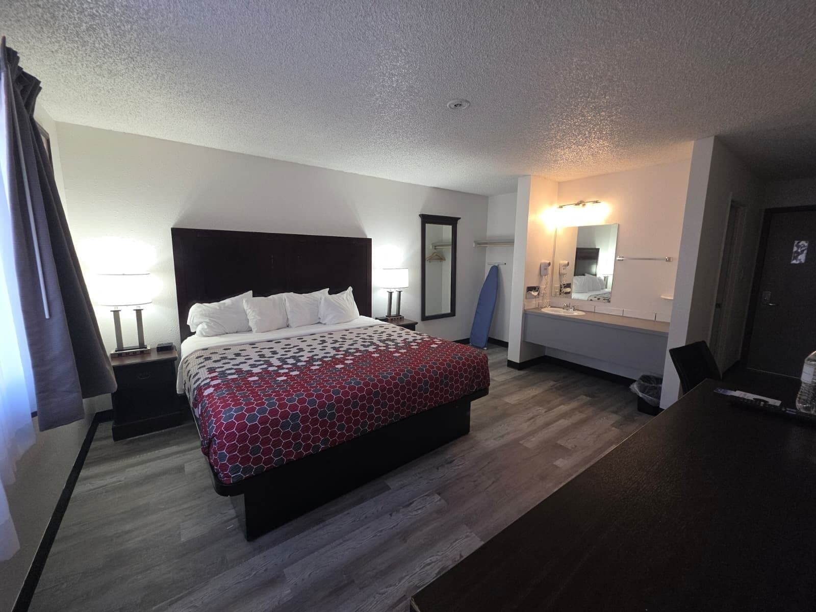 A hotel room with a bed, bedside table, lamp, mirror, and a small desk with a chair.