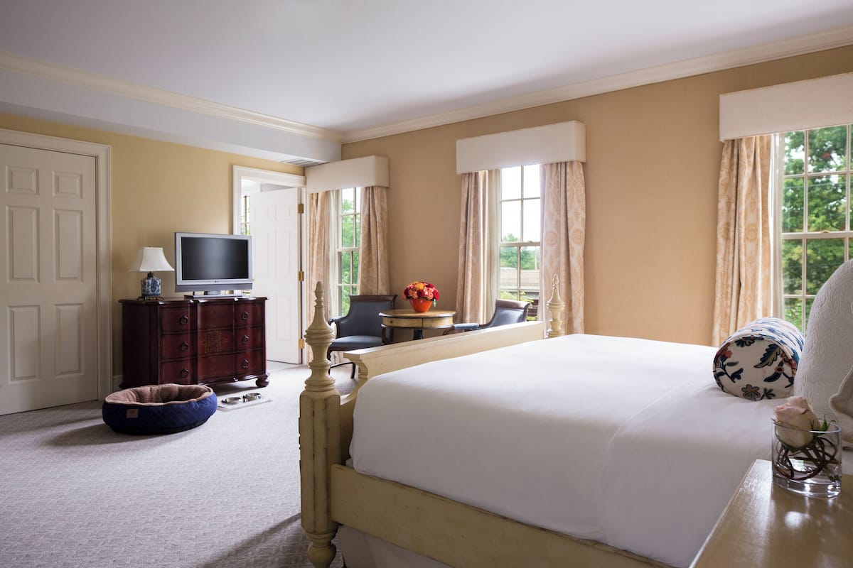 A hotel room with a large bed, a television, a desk with a vase of flowers, and a window with curtains.